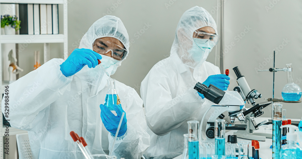 Two Male Scientists in PPE Suit working in Lab while Checking Result of Blood Sample testing. Blue Tone. SARS-CoV-2 , Covid-19 THEME. Two Male Scientists in PPE Suit working in Lab while Checking Result of Blood Sample testing. Blue Tone. SARS-CoV-2 , Covid-19 THEME.