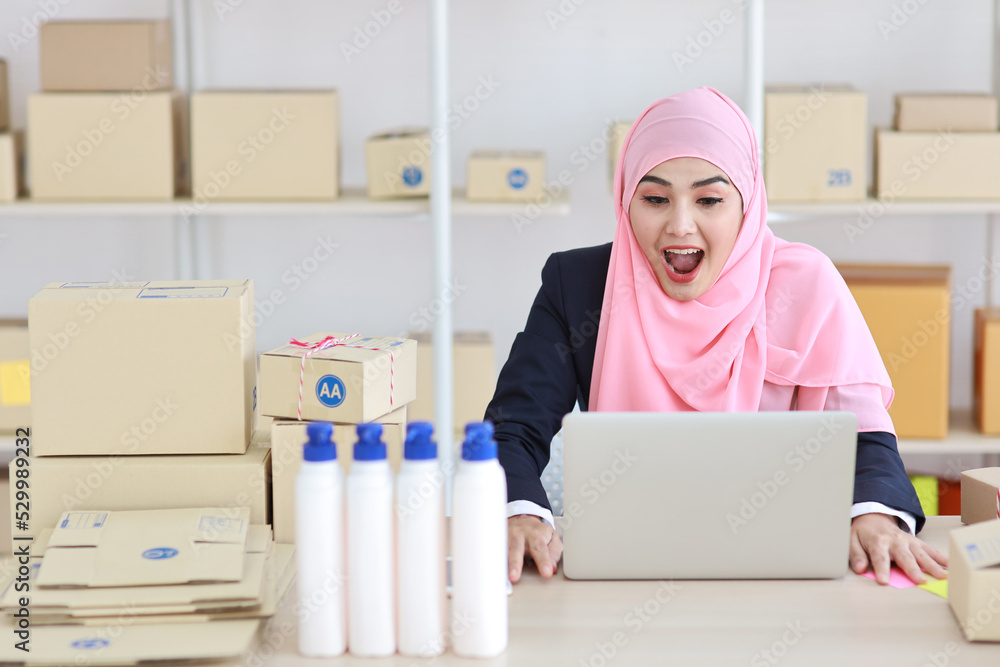 Active asian woman in blue muslim suit sitting and working with computer and online package box delivery. Startup small business freelance working girl look at camera with exciting emotion. Active asian woman in blue muslim suit sitting and working with computer and online package box delivery. Startup small business freelance working girl look at camera with exciting emotion.