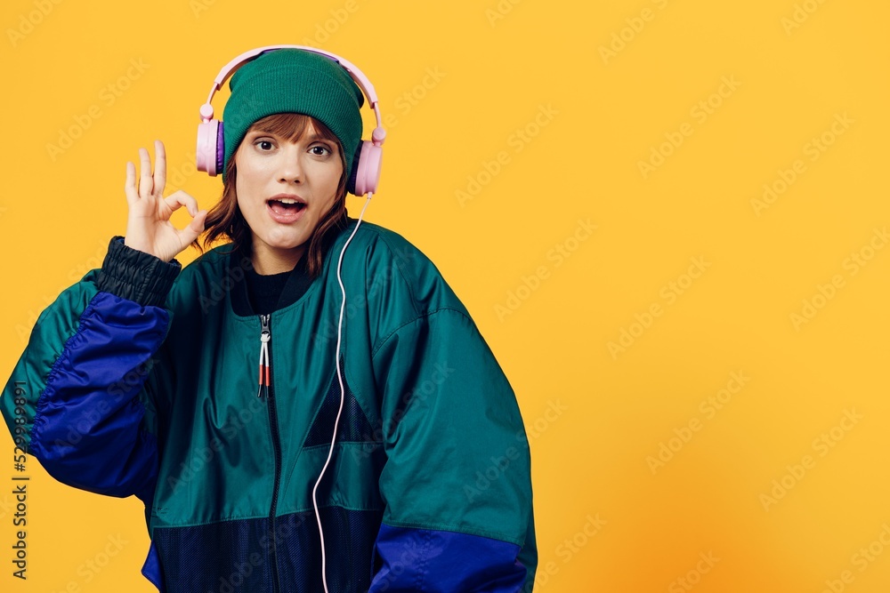 a joyful woman in a green jacket and a hat in the style of the 90s enjoys music standing in pink headphones on a yellow background showing the ok sign while looking at the camera a joyful woman in a green jacket and a hat in the style of the 90s enjoys music standing in pink headphones on a yellow background showing the ok sign while looking at the camera
