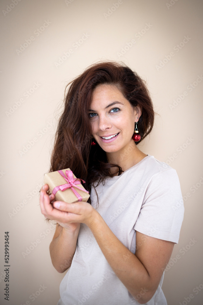 pretty young woman with beautiful long hair is holding a small christmas gift in her hand and has earrings that look like christmas tree baubles standing in front of light brown background pretty young woman with beautiful long hair is holding a small christmas gift in her hand and has earrings that look like christmas tree baubles standing in front of light brown background