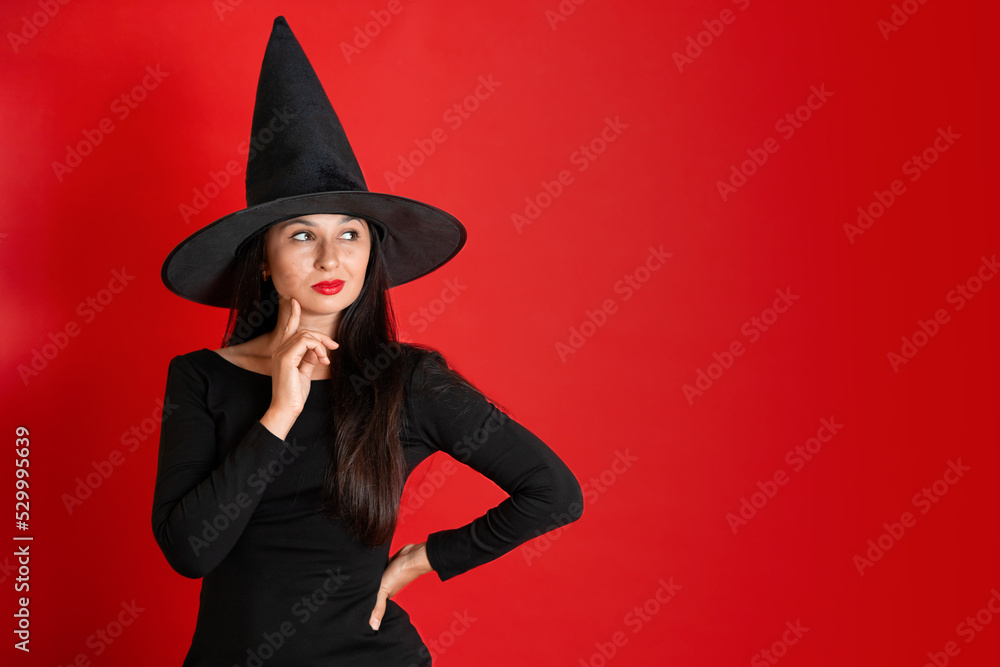 Halloween. A young beautiful woman in a witch's hat and a black dress on a red colored background. Space for text. Halloween. A young beautiful woman in a witch's hat and a black dress on a red colored background. Space for text.