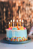 Colorful birthday cake with sprinkles and burning candles on the sparkling gold tinsel background. Festive birthday celebration, party. Vertical card. Selective focus, copy space