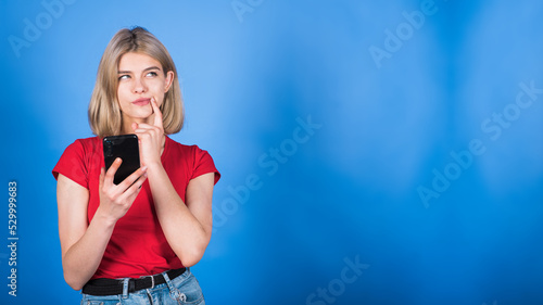 Young caucasian blonde girl in casual clothes holding a finger to her lips, using a mobile phone, thinking and looking at a copy space for advertising isolated on a blue studio background.