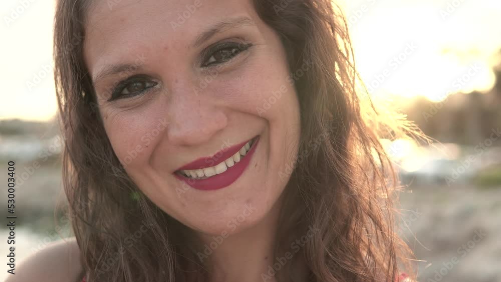 Brunette woman with curly hair and red top looking down and then looking at camera smiling at sunset by the sea. Short shot video of a young girl with a beautiful smile smiling as the camera zooms in. Brunette woman with curly hair and red top looking down and then looking at camera smiling at sunset by the sea. Short shot video of a young girl with a beautiful smile smiling as the camera zooms in.