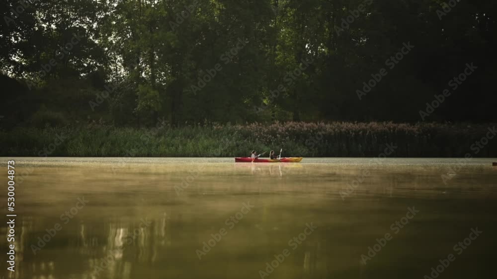 Two people are paddling a kayak in a forest lake at sunrise. Two women are traveling and kayaking in a canoe in a beautiful forest, enjoying vacation together. A morning lake over which fog is creepin Two people are paddling a kayak in a forest lake at sunrise. Two women are traveling and kayaking in a canoe in a beautiful forest, enjoying vacation together. A morning lake over which fog is creepin