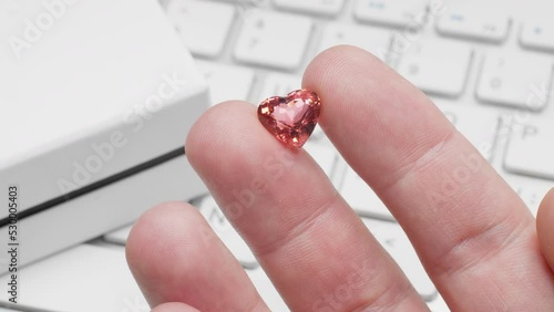 Heart shaped tourmaline gem in hand of gemologist or jeweller. Online jewelry shopping for birthday, Valentine Day or other holiday. Male hand holding red tourmaline, macro shot with box and keyboard.