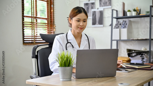 Beautiful Asian female doctor working at her office desk, using laptop computer.