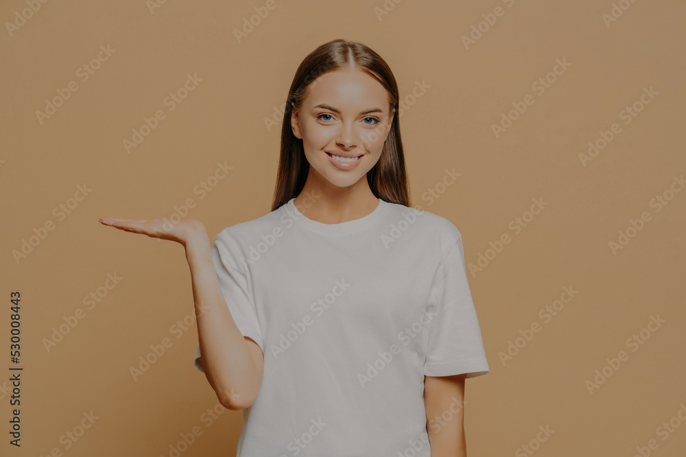 Glad positive young woman raises palm over blank copy space demonstrates invisible object wears casual white t shirt poses against brown background. People advertisement and promotion concept Glad positive young woman raises palm over blank copy space demonstrates invisible object wears casual white t shirt poses against brown background. People advertisement and promotion concept