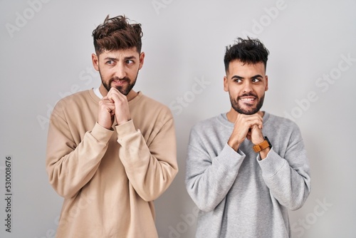 Young homosexual couple standing over white background laughing nervous and excited with hands on chin looking to the side