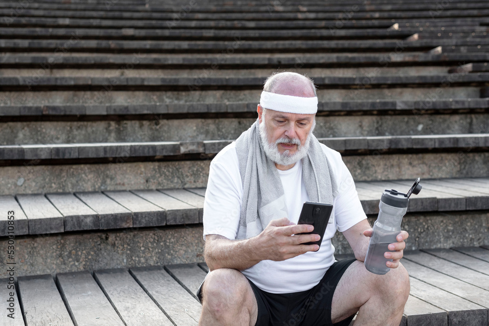 Obraz premium elderly man using smartphone while sitting on urban stairs after training outdoors, resting after fitness workout in city park