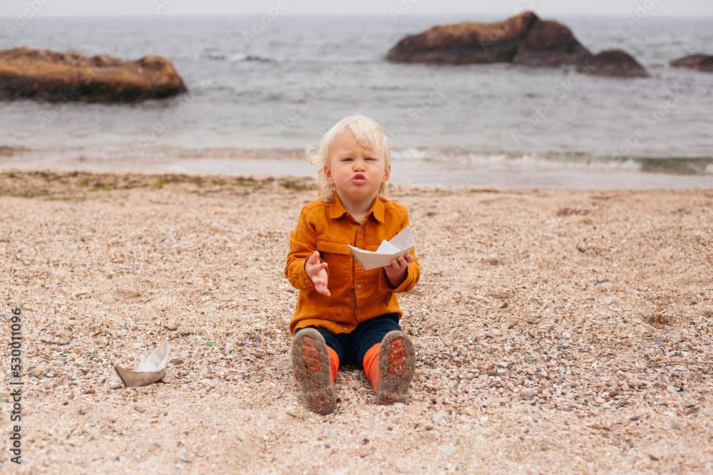 baby boy playing with paper boat on sand on beach in autumn time baby boy playing with paper boat on sand on beach in autumn time