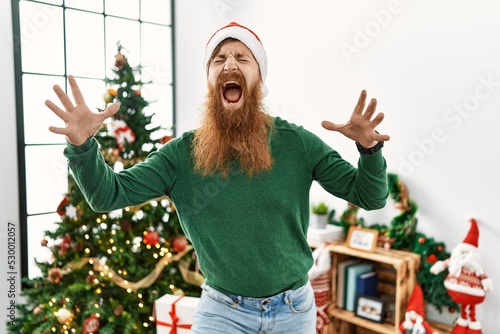Canvas Print Redhead man with long beard wearing christmas hat by christmas tree celebrating mad and crazy for success with arms raised and closed eyes screaming excited