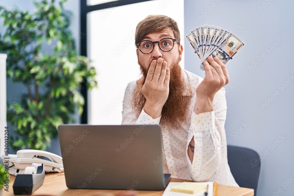 Caucasian man with long beard working using computer laptop holding dollars covering mouth with hand, shocked and afraid for mistake. surprised expression Caucasian man with long beard working using computer laptop holding dollars covering mouth with hand, shocked and afraid for mistake. surprised expression