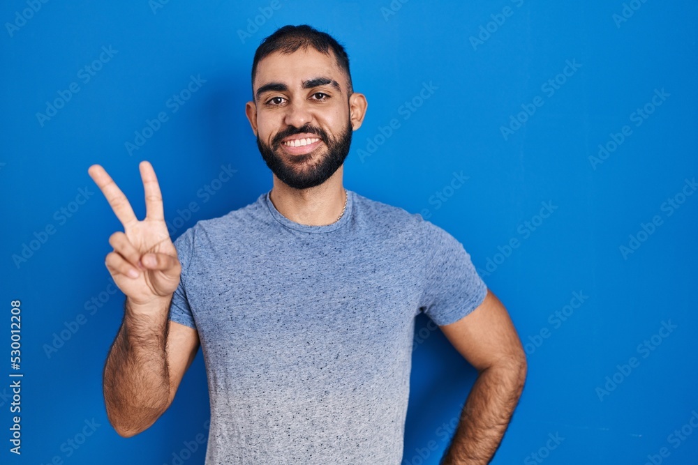 Middle east man with beard standing over blue background smiling looking to the camera showing fingers doing victory sign. number two. Middle east man with beard standing over blue background smiling looking to the camera showing fingers doing victory sign. number two.