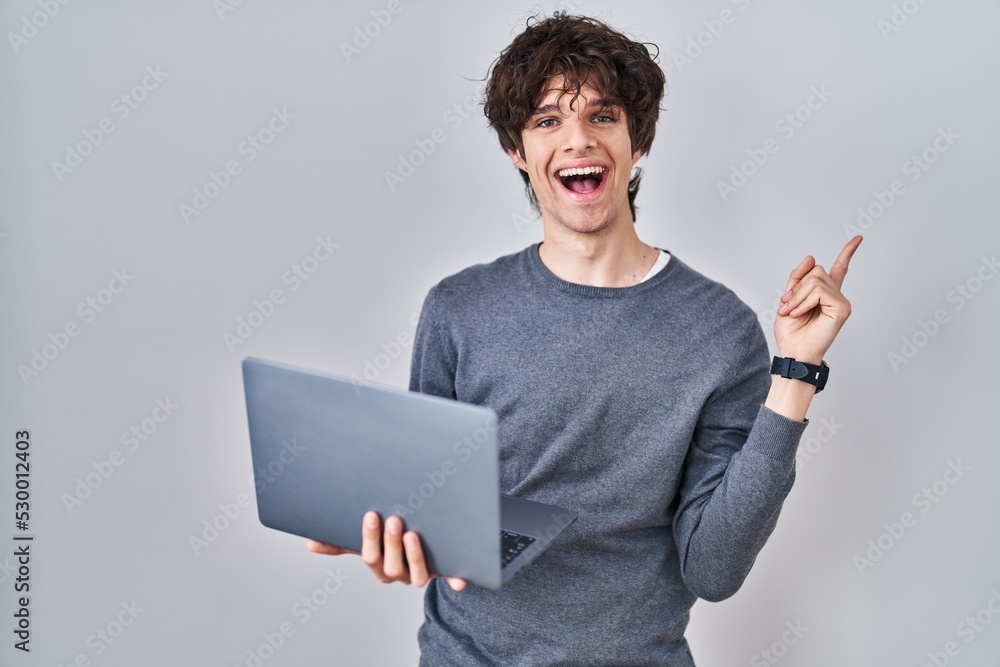 Young man working using computer laptop smiling happy pointing with hand and finger to the side Young man working using computer laptop smiling happy pointing with hand and finger to the side