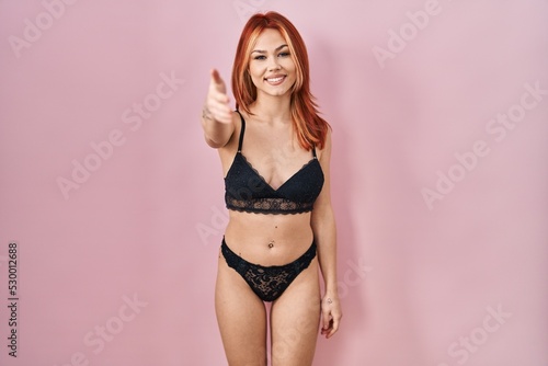 Young caucasian woman wearing lingerie over pink background smiling friendly offering handshake as greeting and welcoming. successful business.
