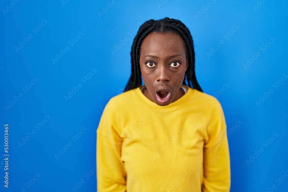 Beautiful black woman standing over blue background afraid and shocked with surprise and amazed expression, fear and excited face. Beautiful black woman standing over blue background afraid and shocked with surprise and amazed expression, fear and excited face.
