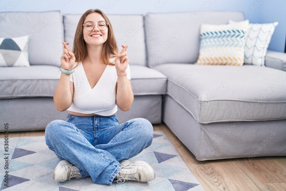 Young caucasian woman sitting on the floor at the living room gesturing finger crossed smiling with hope and eyes closed. luck and superstitious concept.