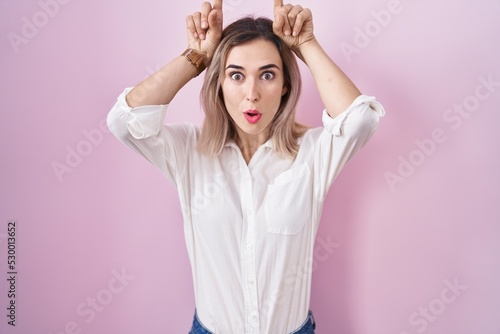 Young beautiful woman standing over pink background doing funny gesture with finger over head as bull horns
