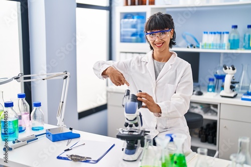 Young hispanic woman working at scientist laboratory smiling happy pointing with hand and finger
