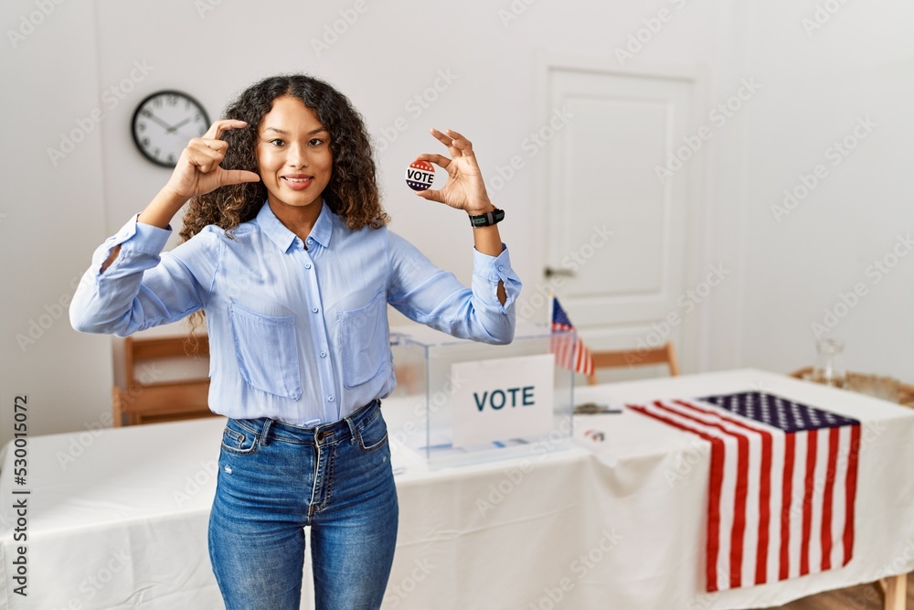 Beautiful hispanic woman standing by at political campaign by voting ballot smiling and confident gesturing with hand doing small size sign with fingers looking and the camera. measure concept. Beautiful hispanic woman standing by at political campaign by voting ballot smiling and confident gesturing with hand doing small size sign with fingers looking and the camera. measure concept.