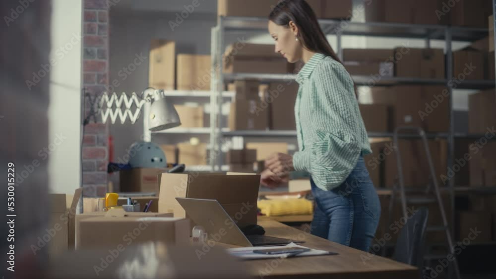 Small Business Owner of an Online Store Works on Laptop Computer while Leaning On Her Desk in Warehouse. Female Employee Packing a Stylish Yellow Jumper in the Room with Shelves Full of Parcels. Small Business Owner of an Online Store Works on Laptop Computer while Leaning On Her Desk in Warehouse. Female Employee Packing a Stylish Yellow Jumper in the Room with Shelves Full of Parcels.