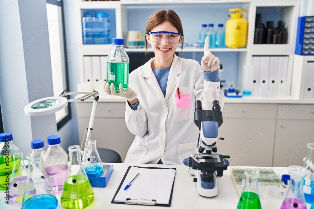 Fototapeta premium Young brunette woman working at scientist laboratory smiling with an idea or question pointing finger with happy face, number one