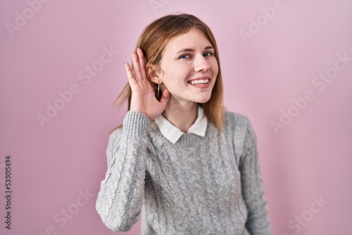 Beautiful woman standing over pink background smiling with hand over ear listening an hearing to rumor or gossip. deafness concept.