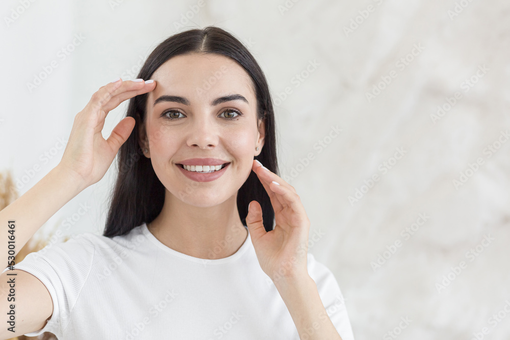 Cosmetology. Close-up portrait of young beautiful brunette woman with clean white skin. He gently touches his face with his hands, looks at the camera, smiles