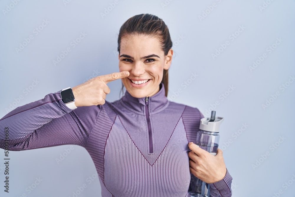 Beautiful woman wearing sportswear holding water bottle pointing with hand finger to face and nose, smiling cheerful. beauty concept Beautiful woman wearing sportswear holding water bottle pointing with hand finger to face and nose, smiling cheerful. beauty concept