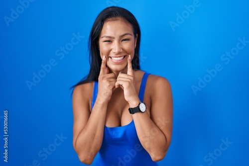 Hispanic woman standing over blue background smiling with open mouth, fingers pointing and forcing cheerful smile photo