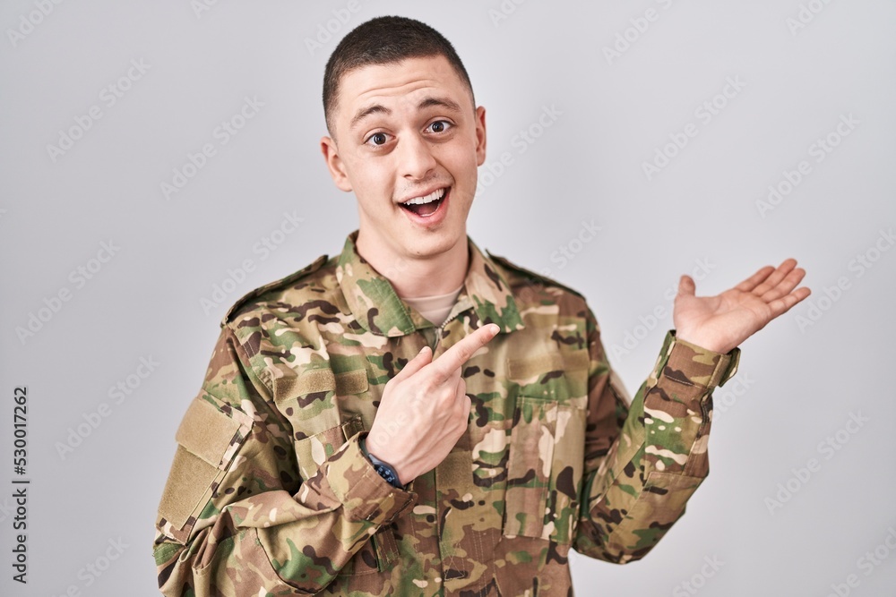 Young man wearing camouflage army uniform amazed and smiling to the camera while presenting with hand and pointing with finger. Young man wearing camouflage army uniform amazed and smiling to the camera while presenting with hand and pointing with finger.