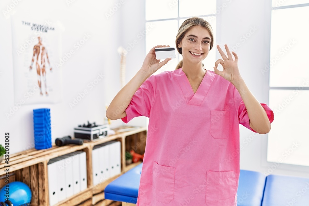 Young physiotherapist woman working at pain recovery clinic holding credit card doing ok sign with fingers, smiling friendly gesturing excellent symbol