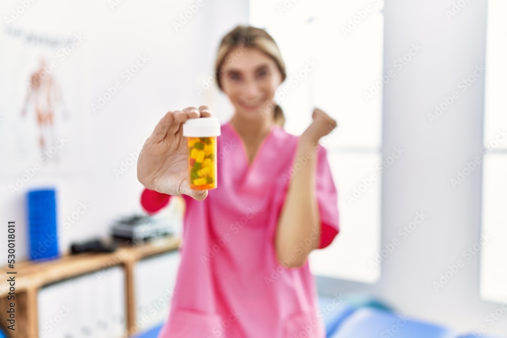 Young physiotherapist woman working at pain recovery clinic holding pills screaming proud, celebrating victory and success very excited with raised arm Young physiotherapist woman working at pain recovery clinic holding pills screaming proud, celebrating victory and success very excited with raised arm