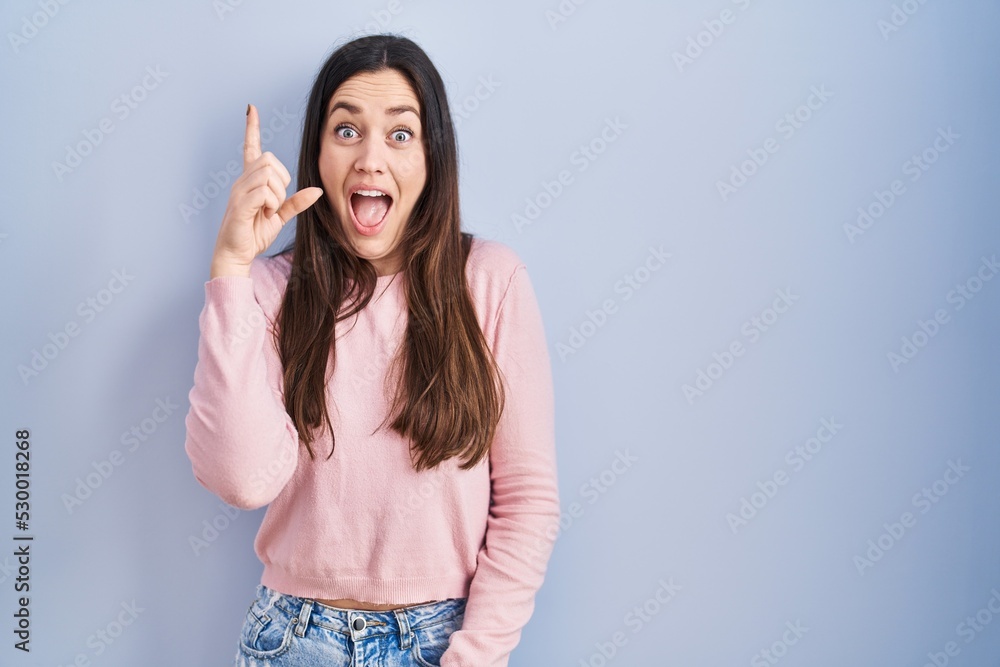 Young brunette woman standing over blue background pointing finger up with successful idea. exited and happy. number one. Young brunette woman standing over blue background pointing finger up with successful idea. exited and happy. number one.