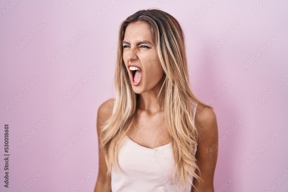 Young blonde woman standing over pink background angry and mad screaming frustrated and furious, shouting with anger. rage and aggressive concept. Young blonde woman standing over pink background angry and mad screaming frustrated and furious, shouting with anger. rage and aggressive concept.