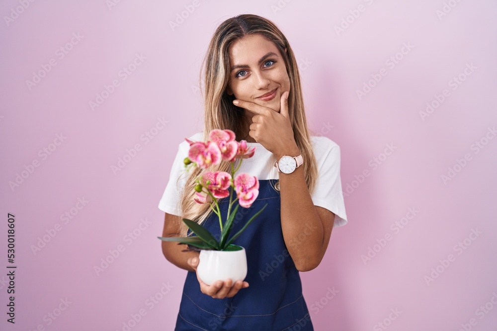 Young blonde woman wearing gardener apron holding plant looking confident at the camera smiling with crossed arms and hand raised on chin. thinking positive. Young blonde woman wearing gardener apron holding plant looking confident at the camera smiling with crossed arms and hand raised on chin. thinking positive.