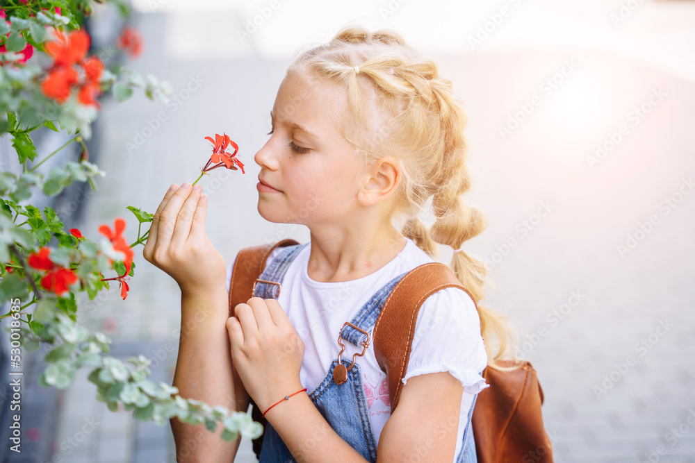 Blond schoolgirl back to school after summer vacations. Child standing with flowers outdoor. Lifestyle portrait of Little caucasian girl with backpack six-seven years old from elementary school.