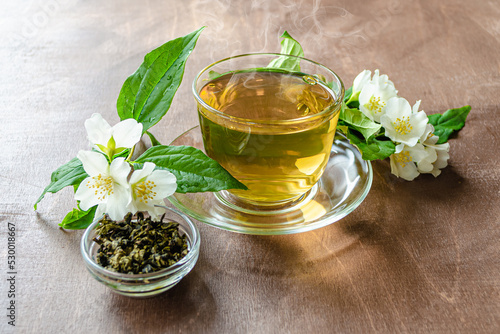 cup of jasmine tea and tea leaves on a wooden table next to jasmine sprigs with leaves and flowers, herbal tea healing diet