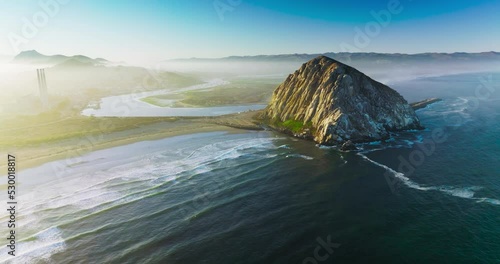Flying closer to a pyramid-shaped mountain at the coast of Pacific. Little white waves coming slowly to the shore. White fog covering the horizon.