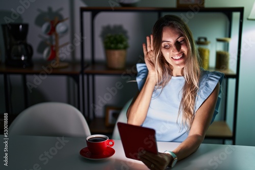 Young hispanic woman using touchpad sitting on the table at night smiling with hand over ear listening an hearing to rumor or gossip. deafness concept.