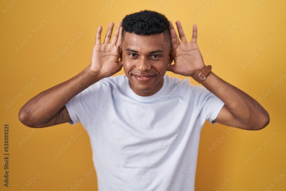 Young hispanic man standing over yellow background trying to hear both hands on ear gesture, curious for gossip. hearing problem, deaf Young hispanic man standing over yellow background trying to hear both hands on ear gesture, curious for gossip. hearing problem, deaf