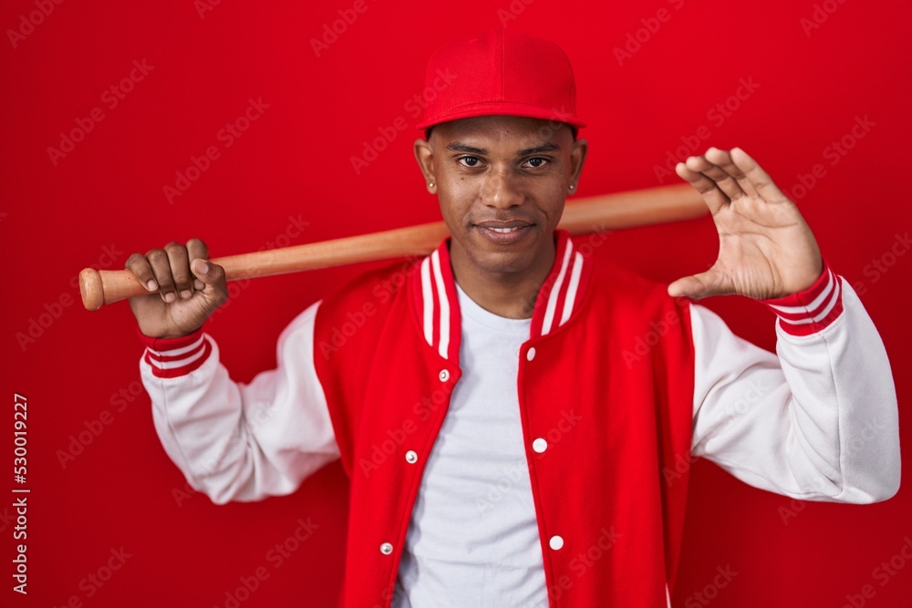 Young hispanic man playing baseball holding bat smiling and confident gesturing with hand doing small size sign with fingers looking and the camera. measure concept. Young hispanic man playing baseball holding bat smiling and confident gesturing with hand doing small size sign with fingers looking and the camera. measure concept.