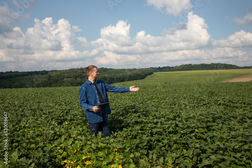 A farmer with a digital tablet checks the quality of soybeans in an agricultural field and points with his hand into the distance. Front view