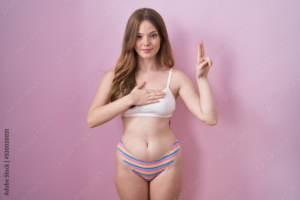 Caucasian woman wearing lingerie over pink background smiling swearing with hand on chest and fingers up, making a loyalty promise oath Caucasian woman wearing lingerie over pink background smiling swearing with hand on chest and fingers up, making a loyalty promise oath
