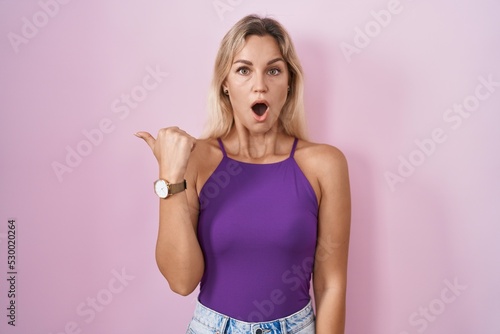 Young blonde woman standing over pink background surprised pointing with hand finger to the side  open mouth amazed expression.
