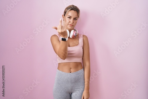 Young blonde woman wearing sportswear and headphones showing middle finger, impolite and rude fuck off expression