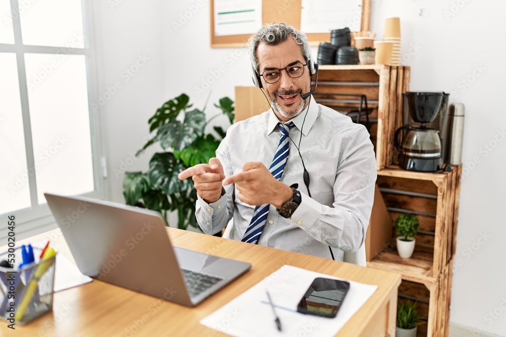 Middle age hispanic business man working at the office wearing operator headset pointing fingers to camera with happy and funny face. good energy and vibes.