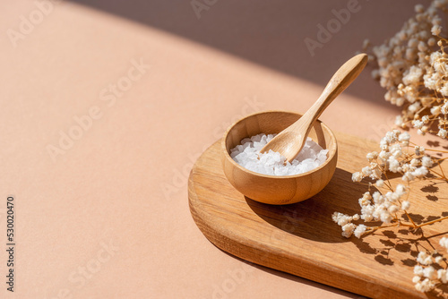 Wooden bowl with a spoon filled with white bath sea salt. Beauty treatment for spa and wellness on beige background. Skincare natural cosmetic concept for body care with dry plants and shadows