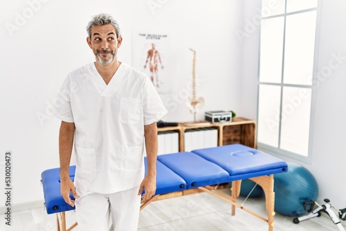 Middle age hispanic therapist man working at pain recovery clinic smiling looking to the side and staring away thinking.